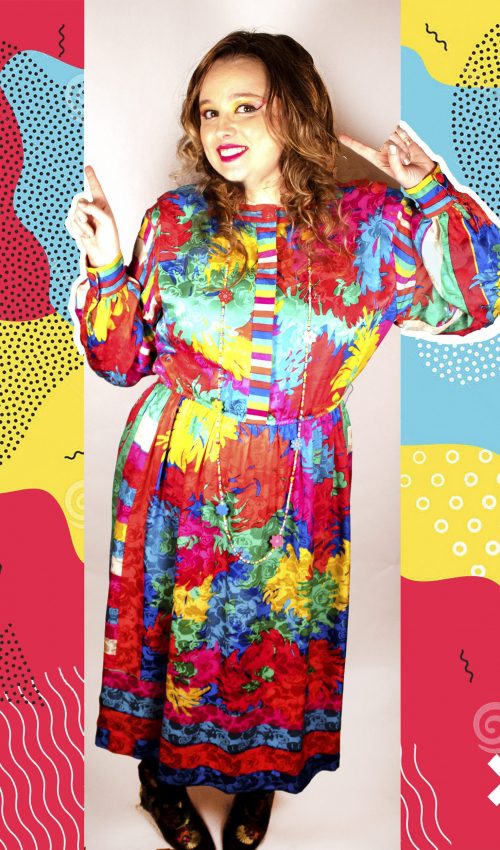Left facing full body photo of Artist Chelsea Smith smiling while looking at us and pointing with both hands to the left. Chelsea is wearing a brightly colored dress and makeup along with a bright 80s inspired design background